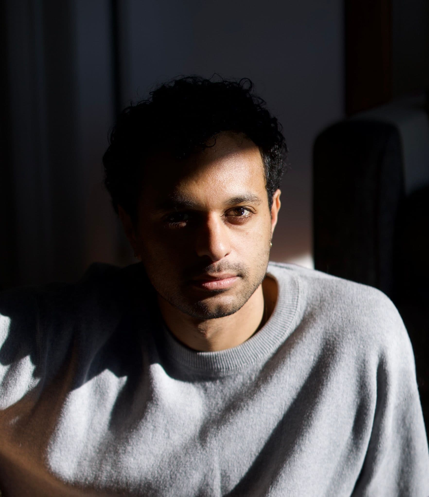 Omar Etman lit by a shaft of window light against a dark room, in a grey sweater.